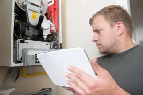 A man inspects a heating system while holding a tablet in a utility room.